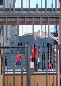 Entrada escolar Carmelites amb reixa arquitectònica, nens jugant a bàsquet al pati.