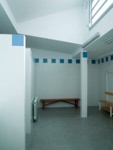 Interior de vestuari amb bancs de fusta, rajoles blanques i detalls blaus.